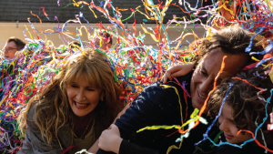 People laughing and hugging while covered in colorful streamers. From L to R: Laura Dern, Will Arnett, and Calvin Knegten in IS THIS THING ON?