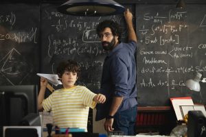 Teacher writing Maths on a chalkboard turning around to see a boy in a yellow striped tshirt throwing a paper plane towards the class