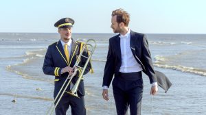 Two men walk along the seashore in formal band attire. The man on the left wears a dark marching band uniform with yellow trim, a cap, and carries a trombone. The man on the right wears a black tuxedo with tails and a white bow tie. They appear to be engaged in conversation, with the ocean waves in the background.