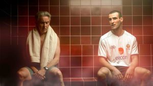 Two men sit on a tiled bench in a steam room. The man of the left, Mick McCarthy (Steve Coogan) has a towel draped over his shoulder. The man on the right Roy Keane (Éanna Hardwicke) wears a white Ireland soccer jersey and dark shorts.