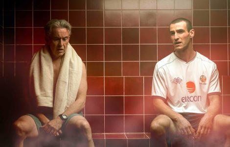 Two men sit on a tiled bench in a steam room. The man of the left, Mick McCarthy (Steve Coogan) has a towel draped over his shoulder. The man on the right Roy Keane (Éanna Hardwicke) wears a white Ireland soccer jersey and dark shorts.
