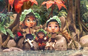 Animated scene of two children and a baby orangutan sitting on rocks beside a stream in a jungle. The children hold large green leaves over their heads like umbrellas. One child wears a school uniform with a red tie, while the other wears traditional jungle clothing and a basket on their back. The orangutan also holds a leaf above its head. Large trees and vibrant foliage surround them.