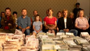 A group of people sits in two rows facing forward, staring intently at large stacks of money piled up in the foreground. The group includes men, women, and a child, dressed in casual clothing, with serious and somber expressions. Behind them is patterned wallpaper and a uniformed guard standing in the background