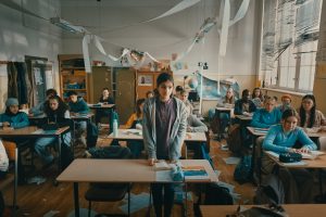Young teenagers all sat in a messy classroom with toilet roll strewn across the roof and classs sheets on the floor. One girl stands awkwardly at the top of the class.