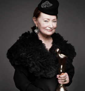 Joan Bergin standing facing camera in black furs holding a staff with a bird head 