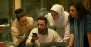 Four people gather closely around a laptop and smartphone in a softly lit office.